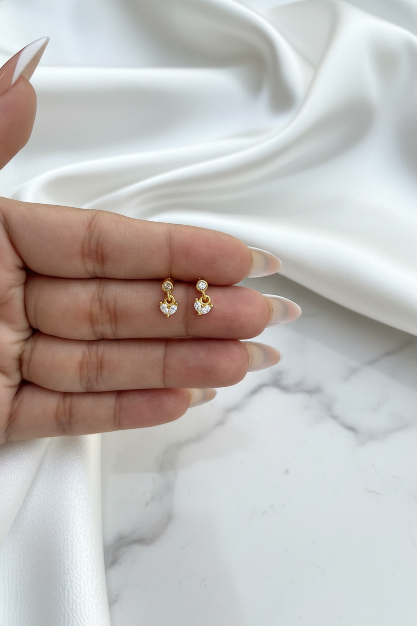 Hand holding gold crystal heart drop stud earrings on white satin marble background