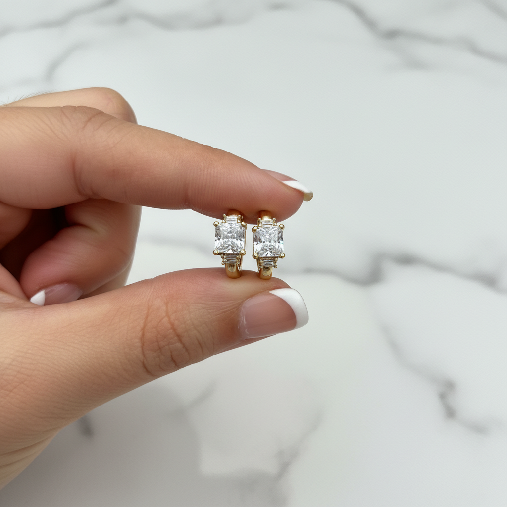 Gold CZ huggie earrings held with French nude nails on white marble